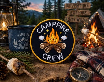 May include: A round, black patch with the words "Campfire Crew" in white, featuring a campfire design with marshmallows. A blue enamel mug with the text "Life is Better Outdoors" is also visible. The scene evokes camping and outdoor adventure.