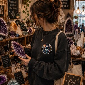 May include: A woman examines a large, raw amethyst crystal in a shop filled with various crystals and gemstones. The shop has wooden shelves and small chalkboard signs. The woman wears a dark gray sweatshirt with a circular graphic and carries a tote bag.