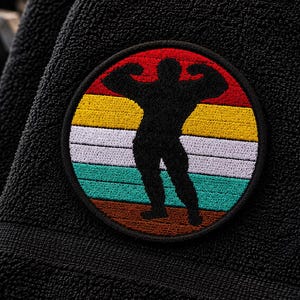 May include: A circular embroidered patch featuring a black silhouette of a bodybuilder flexing muscles against a background of horizontal stripes in red, yellow, white, teal, and brown, stitched onto a black textured fabric towel.