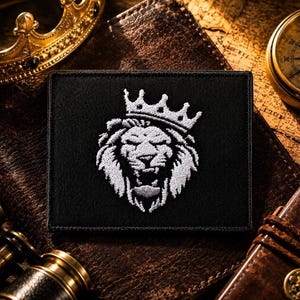 May include: A rectangular black patch featuring a white embroidered lion head wearing a crown. The lion is depicted with a full mane and an open mouth, centered on the fabric. The patch has a stitched border and a flat, textured surface.