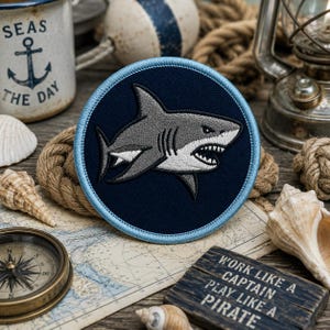 Puede incluir: Un parche redondo azul marino con un borde azul claro que presenta un tiburón gris con dientes blancos. El parche está rodeado de elementos náuticos, como una brújula, conchas marinas, cuerda y una taza que dice "SEAS THE DAY". Un pequeño letrero de madera dice "WORK LIKE A CAPTAIN PLAY LIKE A PIRATE".