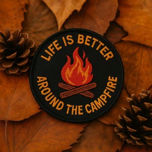 May include: A round black patch with the text "LIFE IS BETTER AROUND THE CAMPFIRE" in orange. The patch features a campfire graphic with orange and red flames above two brown logs. The patch is surrounded by brown leaves and pinecones.