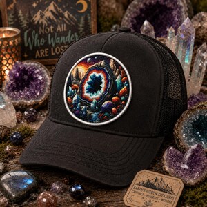 May include: Black trucker hat with a circular patch featuring a colorful geode design. The patch has a white border and depicts a landscape with crystals and a starry sky. The hat has a mesh back and a curved brim.