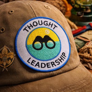 May include: A tan baseball cap featuring a circular embroidered patch with the text THOUGHT LEADERSHIP. The patch displays a yellow sun over a teal horizon with black binoculars in the center. A small gold-toned emblem is visible on the side.