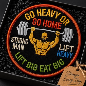 May include: A circular embroidered patch with a black background and orange border. It features a yellow muscular figure lifting a grey barbell. Text reads: GO HEAVY OR GO HOME, STRONG MAN, LIFT HEAVY, LIFT BIG EAT BIG. A brown tag reads Stronger Every Day.