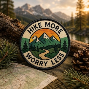 May include: Circular patch with the text "HIKE MORE WORRY LESS" surrounding a mountain scene with a river. The patch is black with a cream-colored border and is set on a wooden surface with a map, pine needles, and a pine cone.