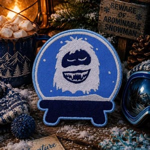 Peut inclure: Un écusson brodé circulaire représentant un yéti blanc avec un large sourire à l'intérieur d'un motif de boule à neige bleue. L'écusson possède une base bleu foncé et est entouré de flocons de neige brodés blancs sur un fond bleu vif.