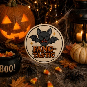 Puede incluir: Un parche bordado circular con fondo beige que presenta un murciélago negro con pajarita roja y el texto naranja FANG-TASTIC. El diseño incluye pequeñas estrellas amarillas y cuenta con un borde cosido alrededor de todo el contorno.