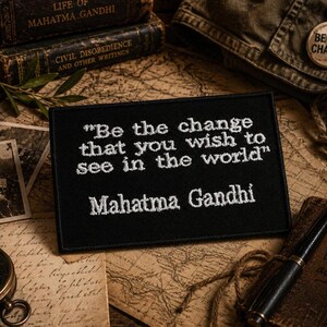Puede incluir: Un parche rectangular negro bordado que presenta la cita en hilo blanco "Be the change that you wish to see in the world" seguida del nombre "Mahatma Gandhi" en una fuente legible, con un borde acabado alrededor del parche.