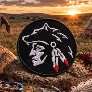 May include: A circular black embroidered patch featuring a white stitched profile of a person wearing a headdress with two feathers. The feathers are detailed with red tips. The patch has a clean, stitched border and a flat, circular shape.