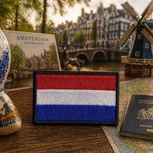 May include: A rectangular patch featuring the Dutch flag with red, white, and blue horizontal stripes. The patch is surrounded by Dutch-themed souvenirs, including a Delftware vase, a wooden clog, and a miniature windmill. A passport and a postcard that says "Amsterdam Netherlands" are also present.