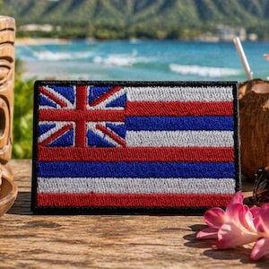 May include: A rectangular embroidered patch featuring the flag of Hawaii, which consists of eight horizontal stripes of white, red, and blue, with the Union Jack in the upper left corner. The patch has a black stitched border and a textured fabric finish.