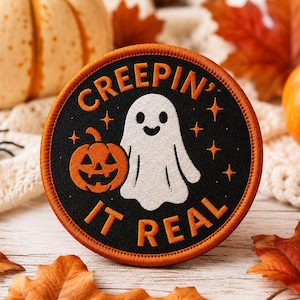 Puede incluir: Un parche redondo negro con borde naranja, con un fantasma blanco y una calabaza naranja. El texto "CREEPIN' IT REAL" es naranja. La imagen está rodeada de calabazas, hojas de otoño y una araña de juguete.