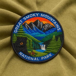 May include: A circular embroidered patch featuring a scenic view of the Great Smoky Mountains National Park. The design includes a black bear, mountains, trees, a waterfall, and the text "Great Smoky Mountains National Park" in a blue border.