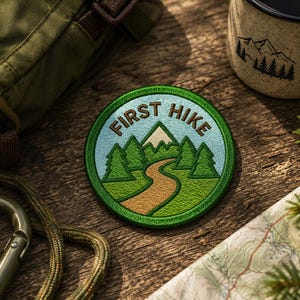 Puede incluir: Un parche bordado circular que muestra un cielo azul claro, un sendero sinuoso de color tostado, pinos verdes y un pico de montaña nevado. El texto "FIRST HIKE" está cosido con hilo marrón a lo largo de la curva superior del parche de borde verde.
