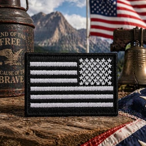 May include: A black and white embroidered American flag patch. The patch features white stars on a black background in the upper left corner and white stripes. The patch is rectangular and has a black border.