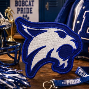 May include: A fabric patch featuring an embroidered white bobcat head silhouette outlined in royal blue. The design is centered, showcasing detailed stitching of the feline facial features, ears, and snout against a dark, textured background.