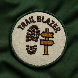 May include: A circular patch with the words "TRAIL BLAZER" in brown lettering. The patch features a brown footprint and a wooden signpost with two directional arrows. The patch is on a dark green background.