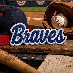 May include: A baseball-themed still life featuring a navy blue and red baseball cap, a baseball glove with a baseball, a wooden baseball bat, and a vintage lineup card. The word "Braves" is embroidered in blue with a white outline.