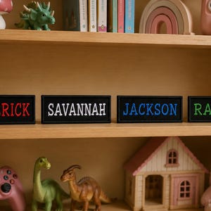 Puede incluir: Cuatro parches rectangulares negros con nombres bordados: "MAVERICK" en rojo, "SAVANNAH" en blanco, "JACKSON" en azul y "RAYLEE" en verde. Los parches se exhiben en una estantería de madera con juguetes y objetos decorativos.