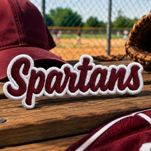 May include: A maroon baseball cap and jersey are next to a baseball glove with a baseball. A maroon and white embroidered patch with the word "Spartans" is on a wooden surface. The background shows a baseball field.