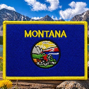 May include: A rectangular embroidered patch with a blue background and a yellow border. The word MONTANA is stitched in yellow letters above a circular emblem featuring a landscape with mountains, a lake, and greenery in white, red, and green.