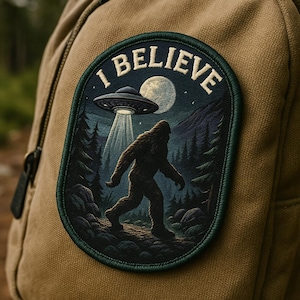 Puede incluir: Parche ovalado con el texto "I BELIEVE" en la parte superior. El parche presenta una silueta de Bigfoot caminando frente a un bosque, con un OVNI que emite un rayo de luz desde el cielo. El fondo incluye una luna llena y montañas.