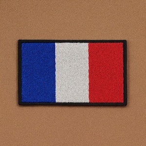Puede incluir: Parche bordado con la bandera francesa, con franjas verticales azules, blancas y rojas, enmarcado por un borde negro. El parche está sobre un fondo de tela marrón.