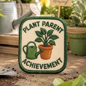 May include: A rectangular embroidered patch with a cream-colored background and a dark green border. The design features a green watering can next to a potted green plant in a terracotta pot, with the text "PLANT PARENT ACHIEVEMENT" in green.