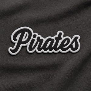 Peut inclure: Écusson brodé avec le mot "Pirates" en écriture cursive. Le texte noir est bordé de blanc, sur un fond en tissu gris foncé. Idéal pour les vêtements ou accessoires.