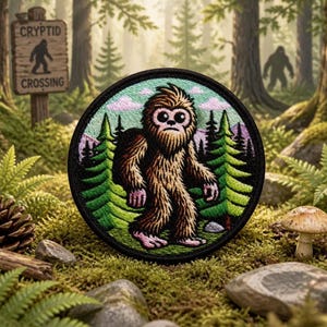 Puede incluir: Un parche bordado circular que presenta una criatura marrón y peluda con ojos grandes de pie en un bosque de pinos verdes. El fondo incluye nubes de color púrpura claro y un borde negro. El diseño representa a un habitante mítico del bosque.