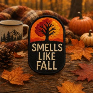 May include: An oval embroidered patch featuring a black silhouette of a tree with red and yellow foliage against an orange sunset background. Below the tree, the text "SMELLS LIKE FALL" is stitched in cream-colored letters on a black background.