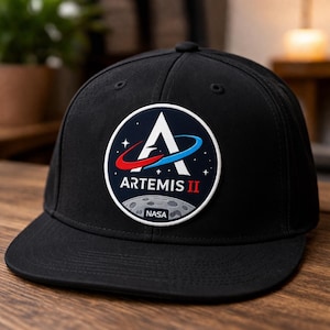 May include: Black baseball cap featuring the Artemis II mission logo. The logo is a white 'A' with red and blue swooshes, stars, and a moon graphic. The text 'ARTEMIS II' and 'NASA' are also visible.