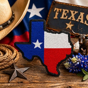 May include: A Texas-shaped patch with the state flag design, featuring blue, white, and red colors. The patch is surrounded by a cowboy hat, rope, a star, a sign that says "TEXAS", a small bronze bull, and bluebonnet flowers, all on a wooden surface.