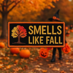 Puede incluir: Parche bordado con el texto "SMELLS LIKE FALL" en hilo dorado sobre fondo negro. El parche presenta un árbol con hojas amarillas y rojas. El parche tiene un borde naranja y está ambientado en una escena otoñal borrosa.