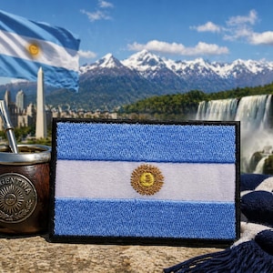 May include: Embroidered patch of the Argentinian flag, featuring light blue and white stripes with a gold sun emblem. A traditional mate cup with a metal straw sits beside the patch. The background includes a waving flag, mountains, and waterfalls.