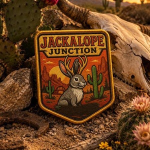 Op de afbeelding: Een rechthoekige geborduurde patch met afgeronde hoeken met een illustratie van een woestijnlandschap. De tekst JACKALOPE JUNCTION staat in vetgedrukte letters boven een grijze jackalope met gewei, omringd door cactussen en een zonsondergang.