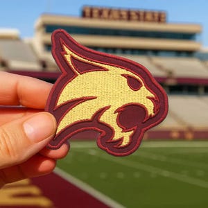 May include: An embroidered patch in maroon and gold, depicting a stylized wildcat head. The patch has a gold interior with maroon detailing. The patch is held up in front of a stadium.