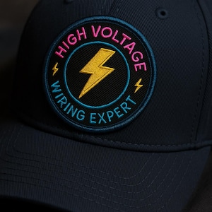 Puede incluir: Gorra de béisbol azul marino con un parche circular. El parche presenta las palabras "HIGH VOLTAGE WIRING EXPERT" en rosa y turquesa, con un rayo amarillo.