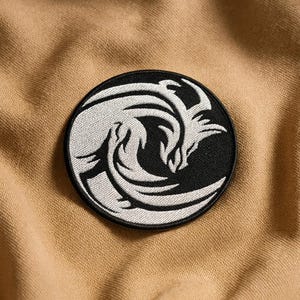 May include: A round, black and white embroidered patch featuring a stylized wave design. The wave is depicted in white thread against a black background. The patch is sewn onto a tan fabric.