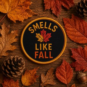 Può includere: Una toppa ricamata circolare con sfondo nero e bordo dorato. Il testo "SMELLS LIKE FALL" è ricamato in lettere dorate e arancioni, centrato attorno a una grafica di foglia d'acero con colori giallo, arancione e rosso.