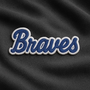 May include: Embroidered patch featuring the word "Braves" in a stylized script. The text is in blue with a white outline, set against a dark gray textured fabric. Ideal for apparel or accessories.