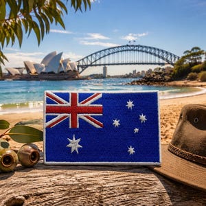 May include: A blue embroidered patch of the Australian flag, with a red and white Union Jack and Southern Cross stars. A brown hat rests on a wooden surface, with Sydney Harbour in the background.