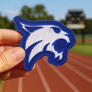 May include: Embroidered patch featuring a white cougar head with blue accents and a blue border. The patch is held up against a blurred background of a track and field. The cougar is a popular mascot for schools and sports teams.