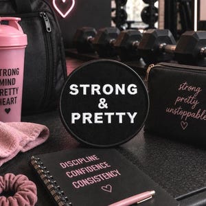 May include: A collection of gym accessories including a pink shaker bottle with the text STRONG MIND PRETTY HEART, a black round patch with white text STRONG & PRETTY, a black pouch with text strong pretty unstoppable, and a black spiral notebook.