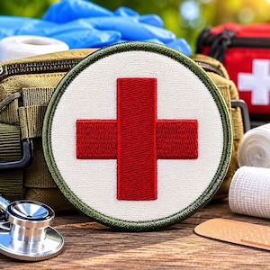 May include: A circular embroidered patch featuring a bold red cross centered on a white background with a dark olive green border. The patch is positioned in front of a tan tactical medical pouch, a stethoscope, white bandages, and a red first aid kit.