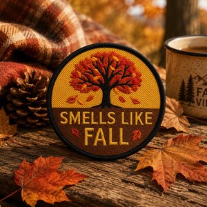 Peut inclure: Un écusson brodé circulaire représentant un arbre noir aux feuilles orange et rouges sur fond jaune, avec le texte "SMELLS LIKE FALL" en lettres jaunes sur fond marron, le tout entouré d'une bordure noire cousue.