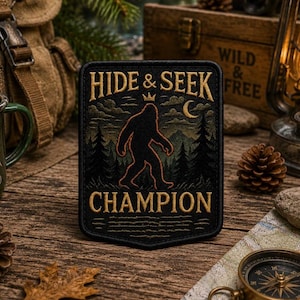 May include: A rectangular black embroidered patch featuring a gold silhouette of a Bigfoot creature walking through a forest. The text HIDE & SEEK is arched above the figure, with the word CHAMPION below, all in gold thread with a small crown icon.
