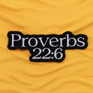 Könnte beinhalten: Ein schwarz-weiß gestickter Aufnäher mit dem Text "Proverbs 22:6" auf gelbem Stoffhintergrund. Der Aufnäher hat einen schwarzen Rand und weiße Buchstaben.