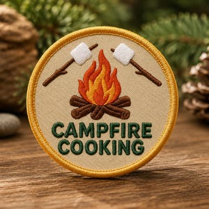 Könnte beinhalten: Ein runder gestickter Aufnäher mit beigem Hintergrund und gelbem Rand. Das Design zeigt ein zentrales Lagerfeuer mit orangefarbenen und roten Flammen über braunen Holzscheiten, flankiert von zwei weißen Marshmallows an Stöcken. Text CAMPFIRE COOKING in Grün.